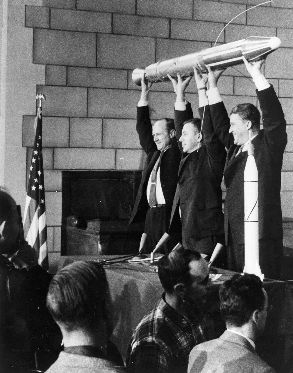 Trois responsables debout tiennent au-dessus de leurs têtes une longue maquette d’Explorer 1, devant des journalistes.